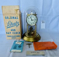 Full view of Schatz 400 day clock with glass dome, brass base, original box, winding key, and manuals spread around.