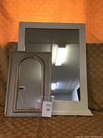 Photo showing two wood framed mirrors set against an orange and brown patterned background. Larger mirror is rectangular with white frame. Smaller mirror is rectangular with arched top and brown wood frame.