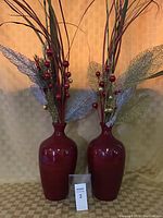 Two red ceramic vases filled with artificial Christmas foliage, featuring long stems, decorative mesh leaves in gold and silver, and red and gold small baubles, on a patterned surface with neutral background.