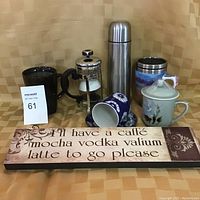 Lot of coffee-related drinkware items on a tan checkered background including French press, thermos, various mugs, and humorous coffee sign