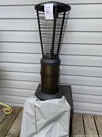 Photo of the full tall patio heater standing on a wooden deck against siding, with a white fabric cover partially draped at the base