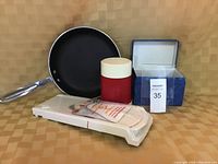 10 inch frying pan, red and white thermos, blue recipe box on a checkered surface photographed together