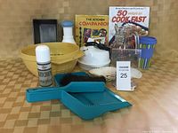 Photo shows assortment of kitchen supplies including bakeware, cookbooks, small kettle, kitchen spray bottle, dustpan with brush, shell-shaped dishes, glass jar, and reusable cup arranged against a plain background.