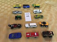 Top-down view of 16 small toy cars arranged in 4 rows on patterned background, showing a variety of vehicle types and colors