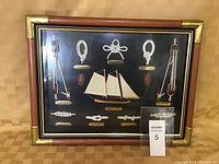 Full view of the nautical shadow box showing multiple knots, wooden pulleys, a miniature sailboat with three sails, all arranged on a black background within a wooden frame with brass corners.