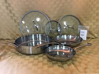 Three stainless steel frying pans with glass lids displayed on a fabric surface, showing sizes and condition.