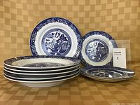 Six blue and white Willow style dinner plates stacked and three side plates laid out, two showing the classic Willow pattern and one a similar decorative blue style plate, all on a brown checkered background.