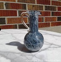 Photo showing front-right angle of the blue and white swirled art glass pitcher vase with clear handle on a white surface in natural light against a brick background.