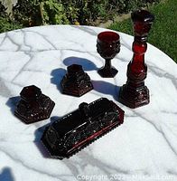 All five Avon Cape Cod ruby red glass items arranged on a white marble surface in outdoor light, showing details and shapes of each piece