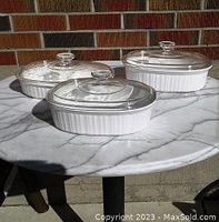 Three white ribbed Corning Ware baking dishes with clear glass lids shown on a marble surface in sunlight.