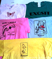 Five t-shirts folded to show their colorful graphic prints: butterfly, EXQSIT text, dolphin with slogan, cartoon tiger, and cartoon duck.