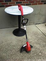 Side view of the red Homelife grass trimmer standing on a concrete surface next to a round white and black table.