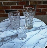 Three heavy cut crystal vases displayed on a marble patterned surface against a brick backdrop, showing the shapes and detailed cuts of each.
