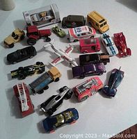 Lot of 18 die-cast toy vehicles arranged on a white surface, showing a range of cars, trucks, an airplane and a boxed car, displaying diverse colors and designs.