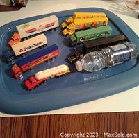 View of 8 assorted toy trucks on a blue tray, showing different designs and colors including yellow, red, white, and green trucks around a water bottle for size reference.