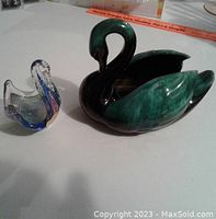 Photo showing both swan figurines together on a white surface, Blue Mountain Pottery swan is larger and darker, glass swan is smaller with purple and blue accents.