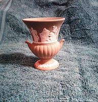 Side angle view of jasperware vase showing classical figures in white relief on muted pink body, ribbed base and small handles