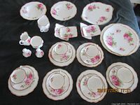Photo showing multiple pieces of Royal Albert American Beauty china including plates, bowls, cups, saucers, small trays, and shakers all with a pink rose floral pattern and gold trim on edges.