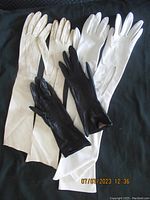 Three pairs of vintage gloves laid flat on black fabric: 2 white pairs (one longer, one shorter) and one black pair (shorter).