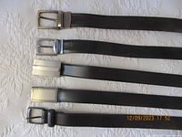 Five men's leather belts arranged flat on a white textured surface. Various buckle styles visible and belts appear in black and dark brown shades.
