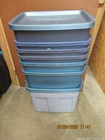 Stack of seven Rubbermaid plastic storage totes with lids. The totes are blue, stacked vertically on a floor surface showing their rectangular shape and uniform sizes.