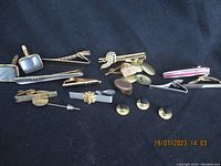 Various men's jewelry items including tie pins, tie bars, and cuff links laid out on dark cloth. Items are vintage and used.