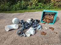 Various plumbing components spread on ground including white PVC pipes, black rubber washers, and copper fittings in a blue crate