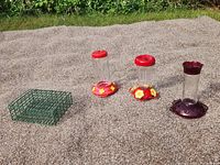 Photo of three hummingbird feeders and a large green metal suet holder placed on a gravel surface outdoors under daylight.