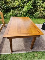 Top and angled side view of medium-toned wooden rectangular dining table on outdoor carpet, showing surface and legs.