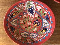 Close-up of one red clay bowl showing detailed floral and bird painted design inside, bright colors with red background.