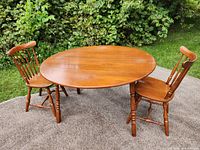 Photo of the vintage round wooden table with two folding leaves, set outdoors on carpet, accompanied by two matching wooden chairs.