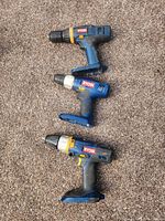 Three Ryobi cordless drills arranged vertically on carpet. Top is a regular drill with yellow ring near chuck, middle is smaller drill with silver around chuck, bottom is a hammer drill with yellow accents near trigger.