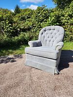 Front and side view of light gray swivel armchair showing tufted backrest, padded armrests, cushioned seat, and skirt covering base.