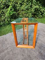 Front and side view of the wood and glass TV stand placed outdoors showing three clear glass shelves and wooden frame with glass top.