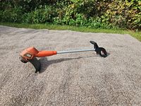 Side view of whole weed wacker showing orange motor housing, long silver shaft, black handle, and guard.