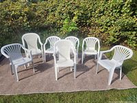 Seven white plastic patio chairs arranged on an outdoor carpet in a garden setting. Chairs have varied design with some having armrests and some not, all white plastic.