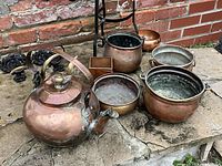 Overview of the collection with copper and brass antiqued pots and kettles in varying sizes and shapes, including a brass and iron stand.