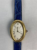 Universal Genève ladies wristwatch laid flat showing oval rolled gold case, silver-tone dial with Roman numerals, and blue textured leather strap.