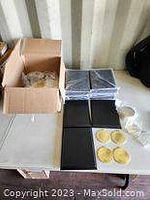 Cardboard box with gold-colored medals in plastic bags, and several black plaques stacked flat on a table