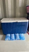 Blue Coleman cooler with white lid and four blue reusable ice packs in front on a surface.
