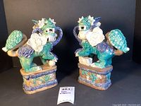 Pair of painted ceramic foo dogs or guardian lions placed side by side showing front and side angles with a label indicating repair.