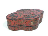 Angled view of red lacquer box showing raised hand painted bat and fruit design on top and side, with some wear visible.