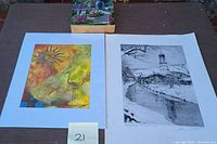Three artworks shown on a surface including a vibrant mixed medium painting with a sun and flamingo, a black and white etching of a snowy village, and an oil painting on wood of a garden scene with elderly person
