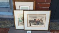 Two framed art prints leaning side by side against a brick wall, one with boats in harbor and one with a covered bridge scene.