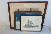 Three framed art prints stacked leaning against a wall, showing edges of mats and frames.