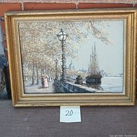 Full view of the framed painting showing a European harbor scene with a tall ship docked, lampposts along a promenade, trees, and figures. The painting is in a gold-tone frame and labeled with '20' on a card below.