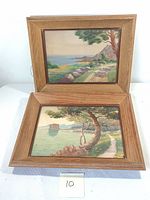 Two framed watercolor paintings showing coastal landscapes with trees and pathways, displayed together on a white surface.