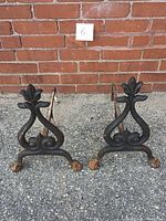 Pair of rustic serpentine figural andirons side by side on gravel in front of brick wall, showing overall form and claw feet.