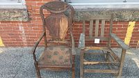 Photo showing both antique chairs side by side against brick wall; rocker chair on left with upholstered seat, wooden arm chair on right in worn condition.