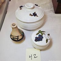 Photo showing two white pottery bowls with lids decorated with raised floral and grape designs, alongside a small Southwestern pottery vase.